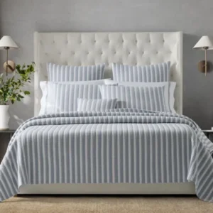 A stylish bedroom featuring a tufted white bed with striped bedding, soft pillows, and modern wall sconces, complemented by a plant. Featuring Apollo bedding in Dove, by Matouk. Available at Linen Society.