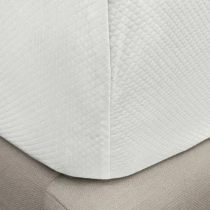 A close-up of a corner of a white quilted bed cover draped over a textured beige mattress, showing fabric details and textures. Featuring the Diamond Piqué by Matouk, available at Linen Society.