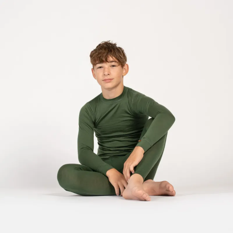 A barefoot child sits comfortably on the floor against a plain white background, wearing a matching dark green long-sleeve top and leggings. One leg is bent with their arm resting on their knee, while they look calmly at the camera.