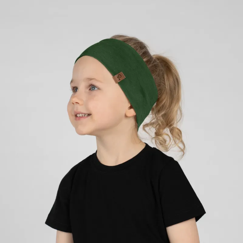 A close-up studio portrait of a smiling young child looking up and to the left against a plain light grey background. She is wearing a black short-sleeved t-shirt and a wide dark green headband with a small brown logo tag on the side, her hair pulled back in a ponytail.