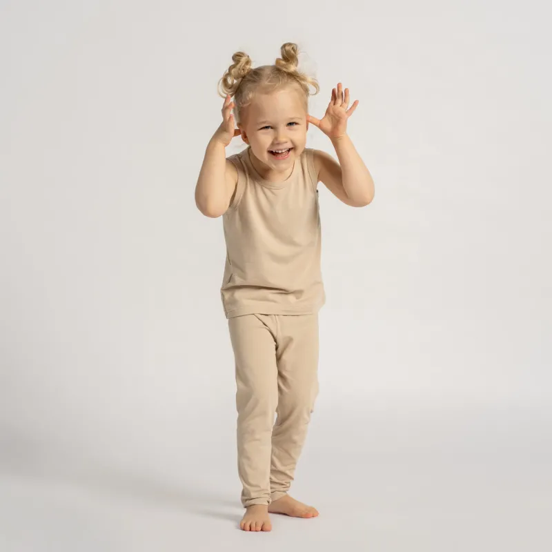 Smiling toddler wearing a beige sleeveless top and leggings set, standing barefoot and playing in front of a light background.