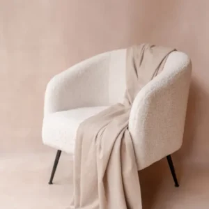 Cashmere Blanket in Oatmeal Melange by TBCo elegantly draped over a textured white armchair with soft fringe detailing