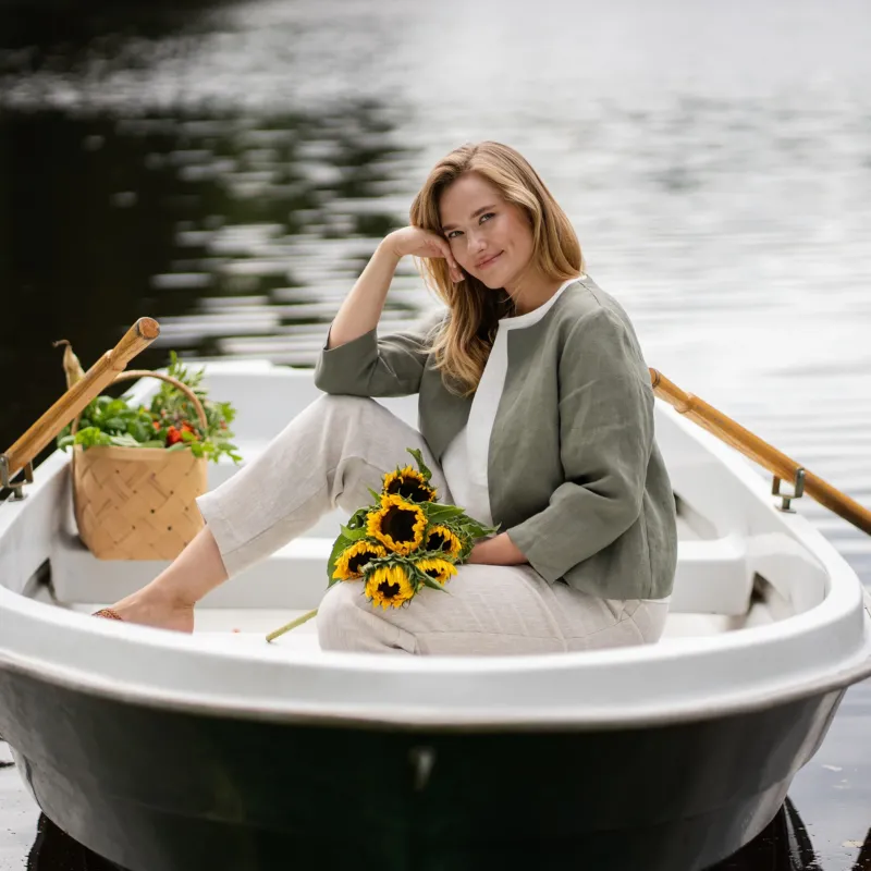 A smiling woman relaxes in a white rowboat on the water, resting her cheek gently against her hand. She is wearing a collarless stone green linen jacket, a white top, and natural linen pants, with a bunch of bright sunflowers resting on her lap.