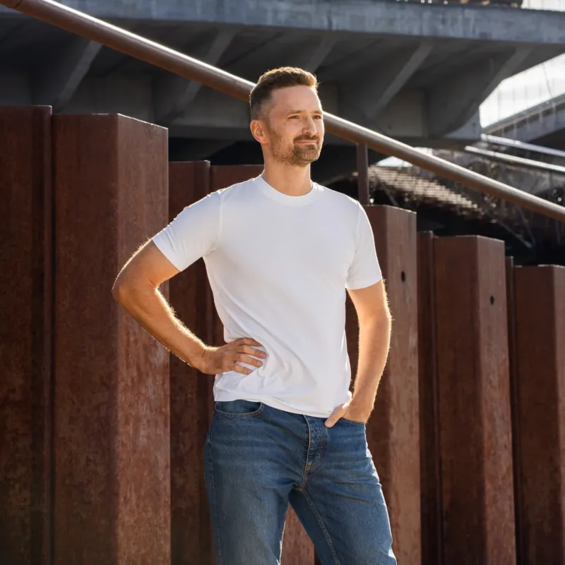 A man stands outdoors against a backdrop of rusted steel beams and a modern concrete overpass. He is wearing a plain white short-sleeved t-shirt and blue jeans. His right hand rests on his hip, and his left hand is tucked into his front pocket as he looks off into the distance.