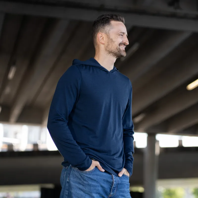 A smiling man stands outdoors with both hands in the front pockets of his blue jeans. He is wearing a long-sleeve, navy blue hoodie. The background shows the underside of a large concrete overpass structure.