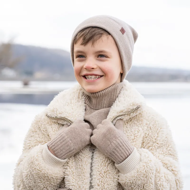 A young child with short brown hair and a wide smile, is dressed in winter clothing. They are wearing a beige beanie, a beige turtleneck sweater, and a cream-colored sherpa jacket, with beige knitted gloves. A snowy, blurred background with what appears to be a body of water is visible behind them.